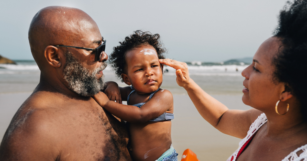 Man holding young girl on beach. Woman applying sunscreen to girl's face